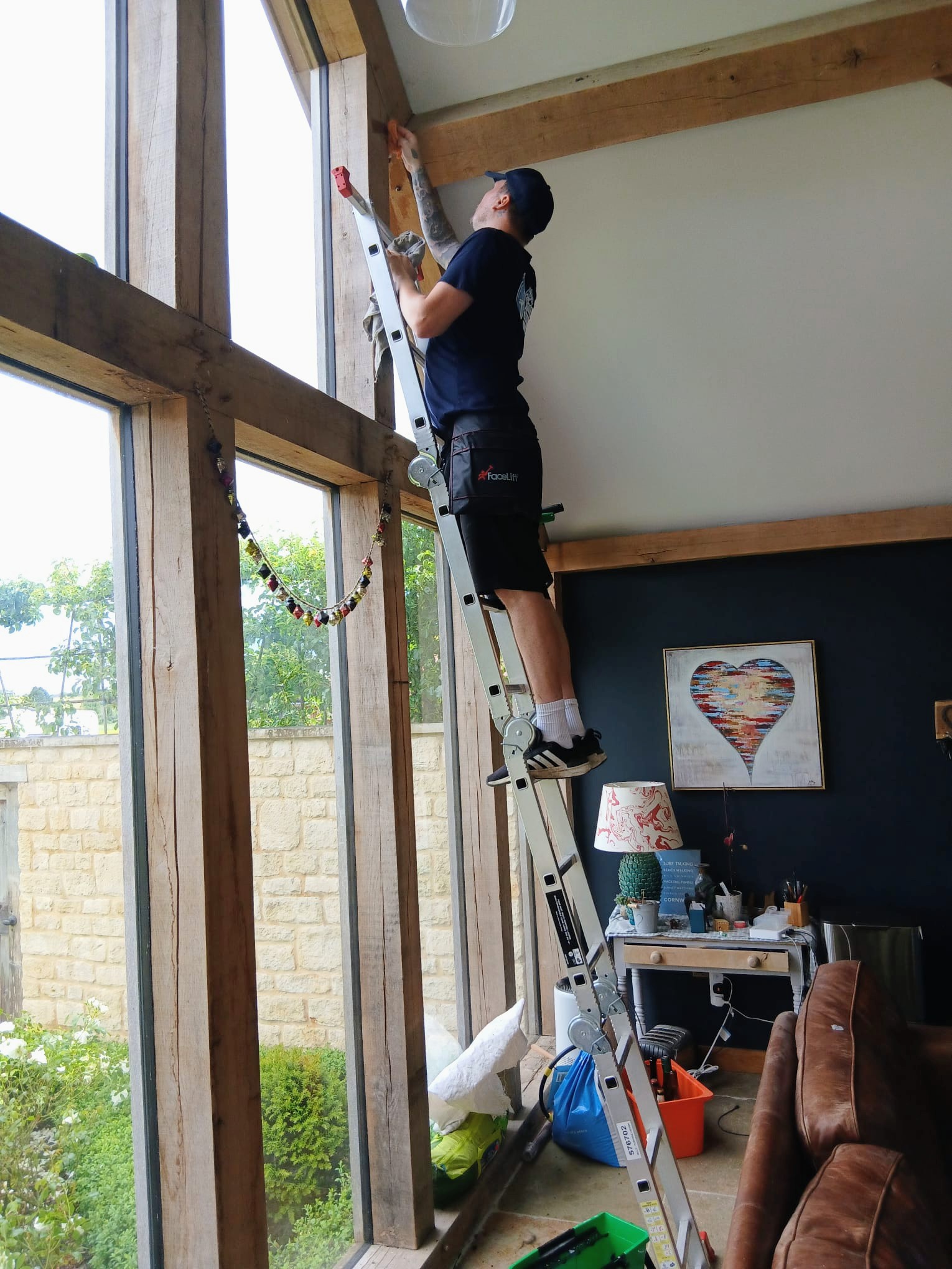 Cleaning tall oak-framed windows from a stepladder inside a high-ceilinged room