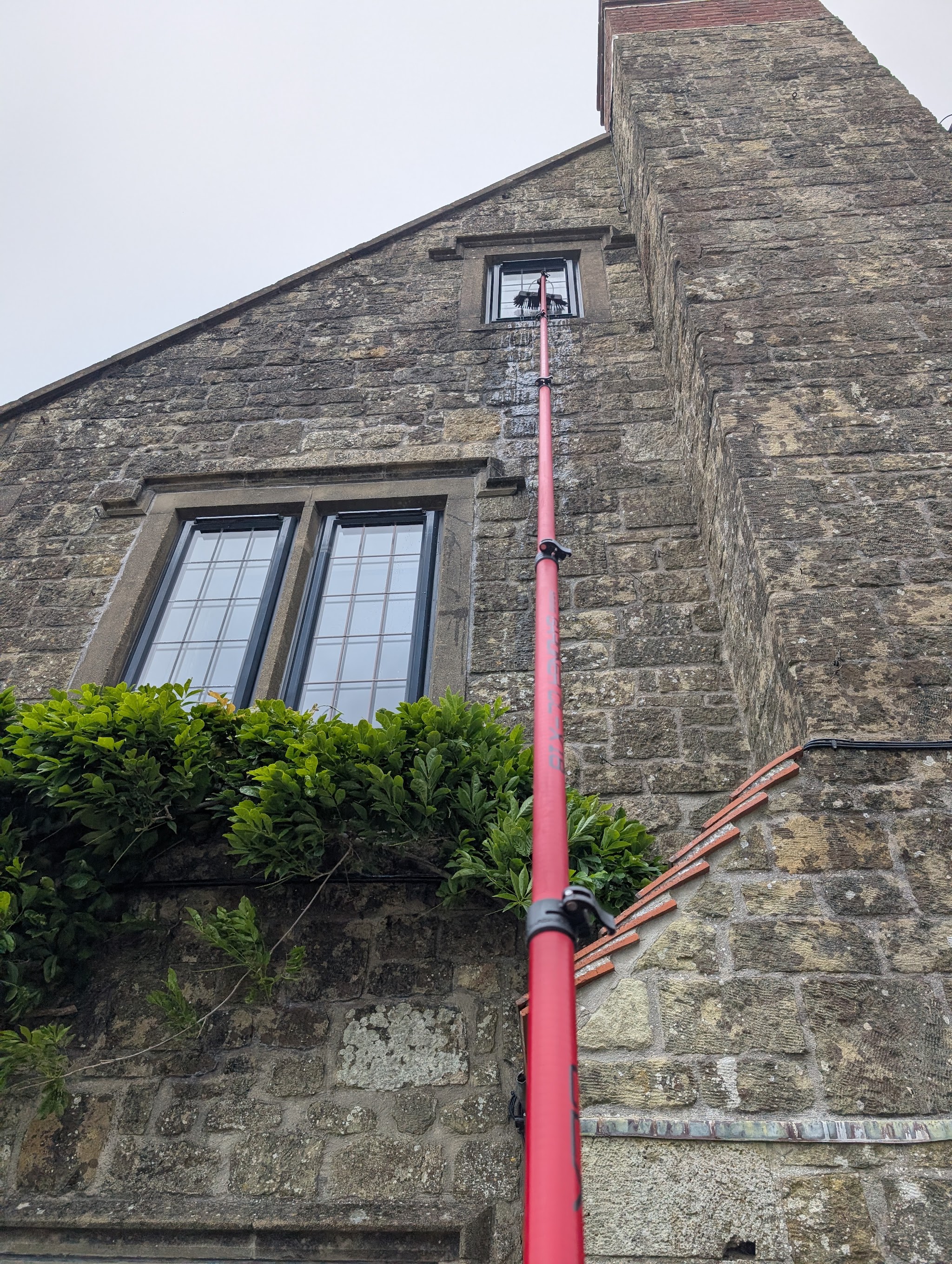 A red water-fed pole reaching an upstairs leaded window on an old stone house